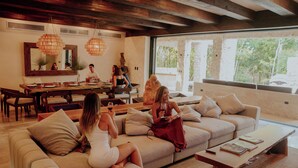Una sala de estar amplia con un sofá grande, una mesa de comedor y una mesa de centro. Varias personas están sentadas y de pie alrededor de la sala.