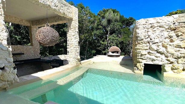 Un área de piscina con paredes de piedra, una esfera de mimbre colgante, una parrilla y una silla de descanso.