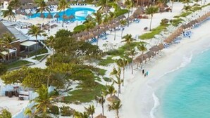 Un resort en la playa con una hilera de sombrillas de paja y sillones de descanso a lo largo de la costa, un área de piscina y un edificio con varios balcones.