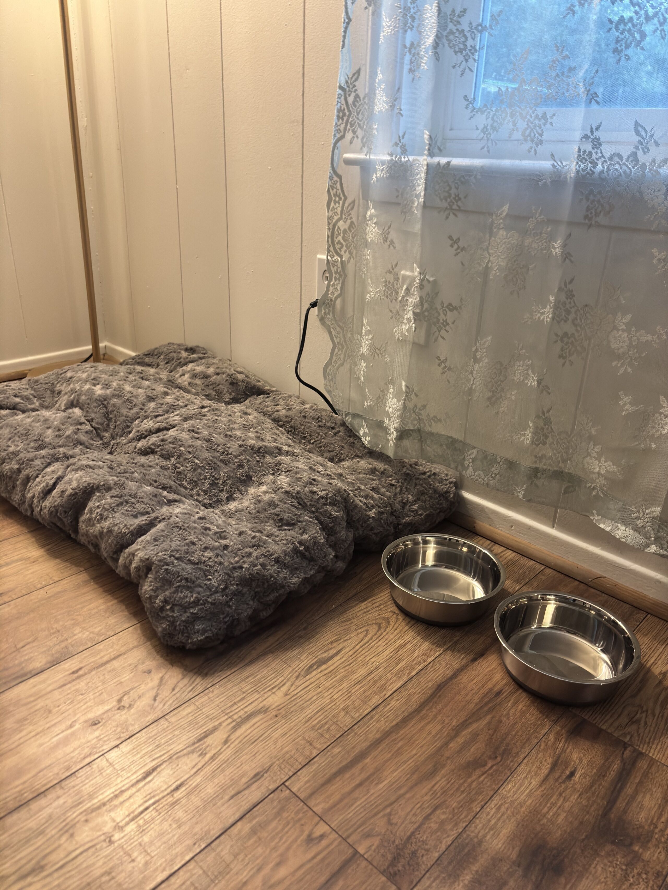 Een dierenbed en twee metalen kommen op een houten vloer, nabij een raam met een dunne gordijn.