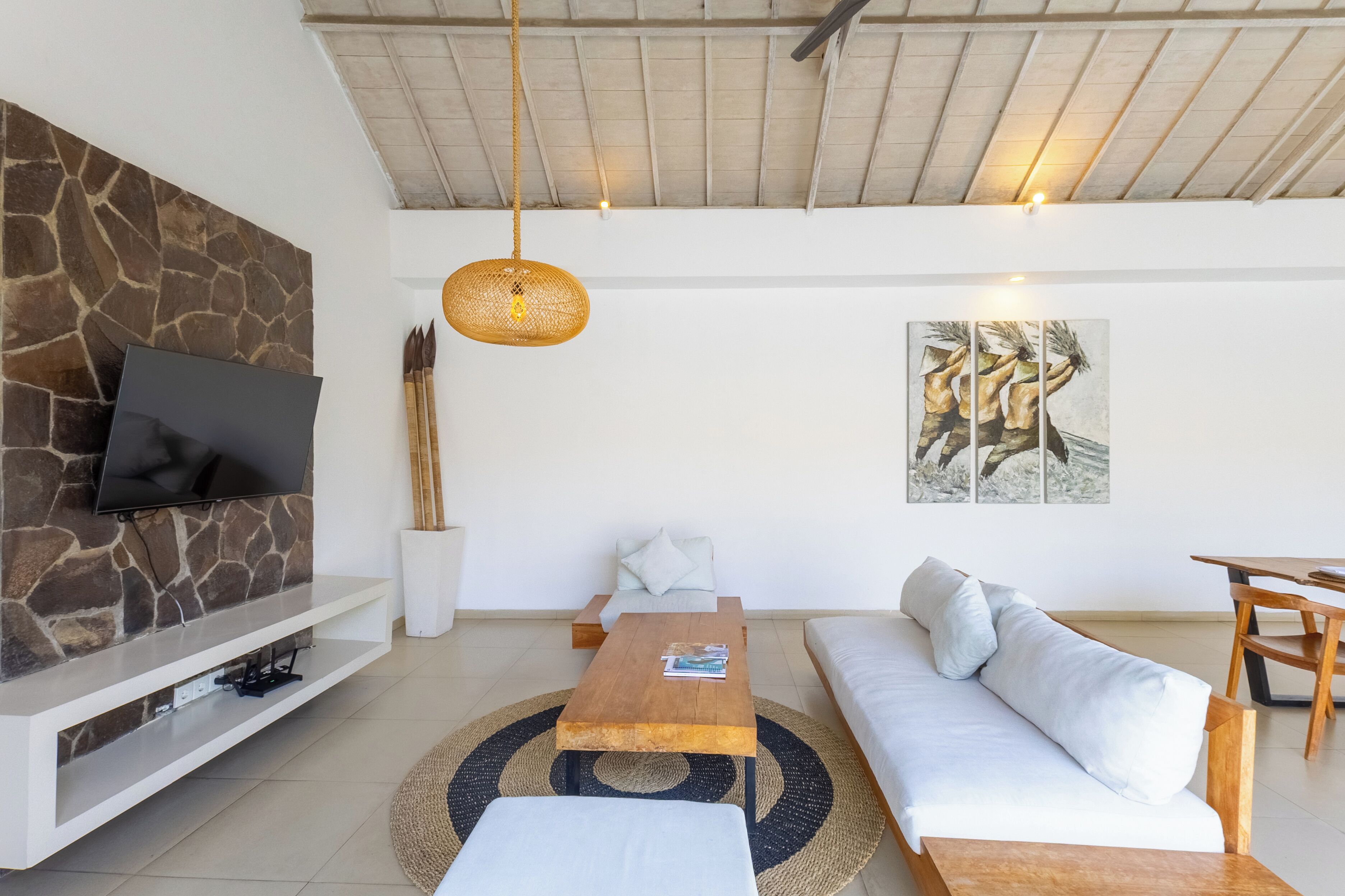 A modern living room with a stone wall, flat-screen TV, wooden coffee table, white sofa, and a dining area with a wooden table and chairs.