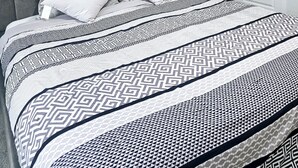 Una cama bien hecha con un cabecero tapizado gris, ropa de cama blanca con estampado y una puerta blanca al fondo.