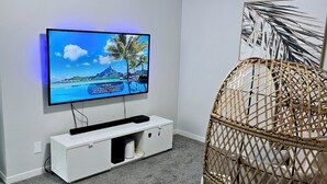 Una habitación con una televisión de pantalla plana montada sobre un mueble blanco, una lámpara de techo, una silla de mimbre y un cuadro enmarcado en la pared.