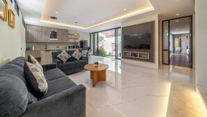 A modern living room with a sofa, coffee table, and TV. There is a kitchen area with cabinets and a dining table. The room has a large window and a door leading to another room. - Villa Noema 2 - Brand New 2BR Villa with Pool (Badung)