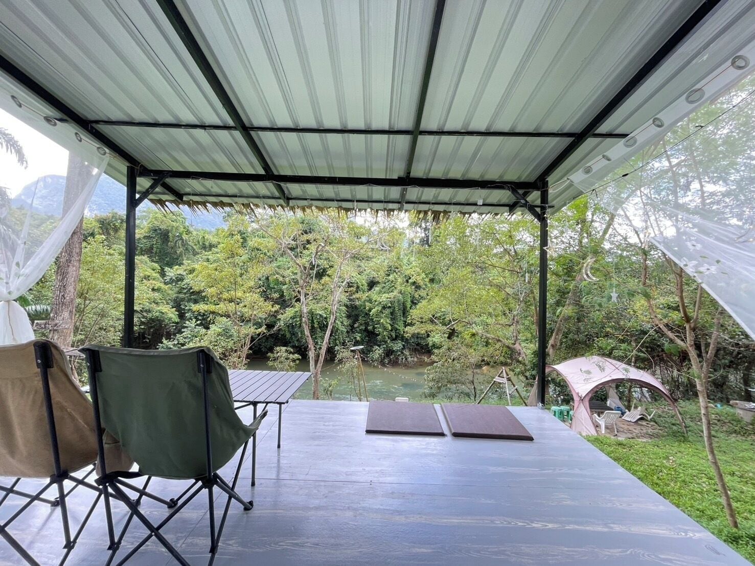 Un espacio al aire libre cubierto con vista a un río y un bosque. Hay sillas plegables y una mesa.