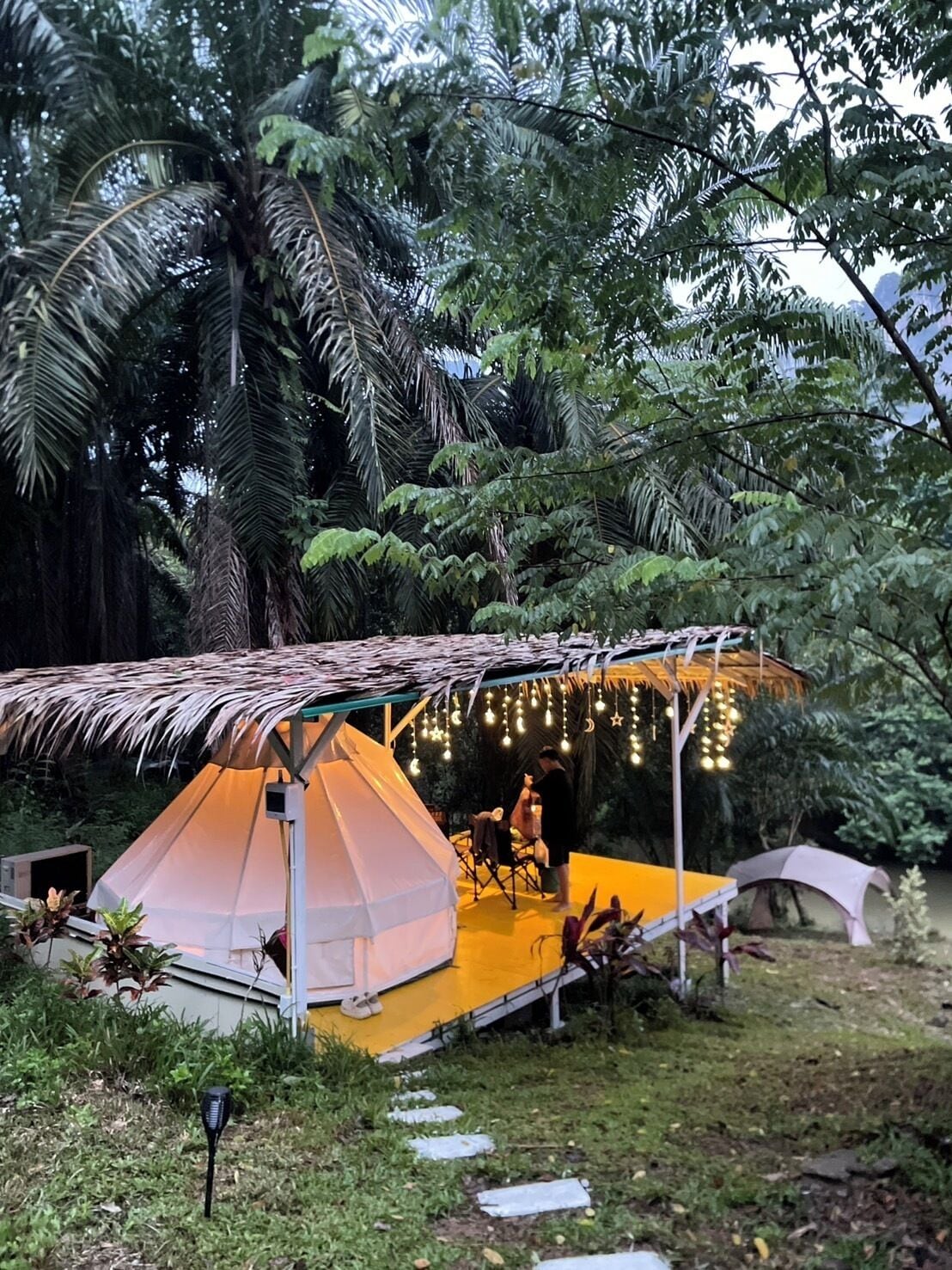Uma área de camping com tendas e um telhado de palha, cercada por vegetação exuberante e palmeiras.