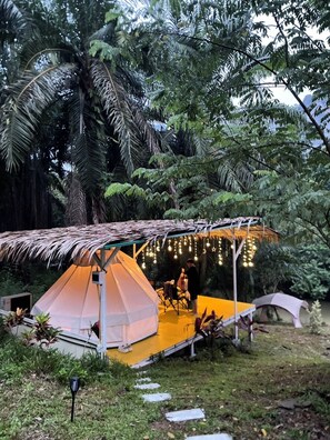 A tented camping area with a thatched roof, surrounded by lush greenery and palm trees. - Amornkeeree Khao Sok Camping (Phanom)