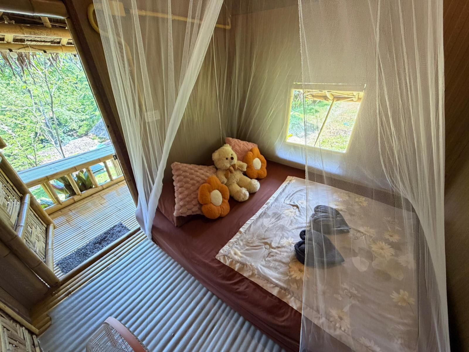 A room with a bed, a window, a mosquito net, and a view of greenery outside.