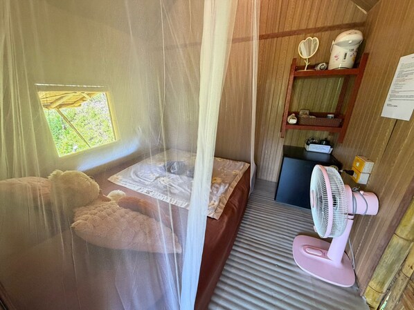 Une chambre avec un lit, un petit bureau, un ventilateur et une fenêtre donnant sur un paysage verdoyant.