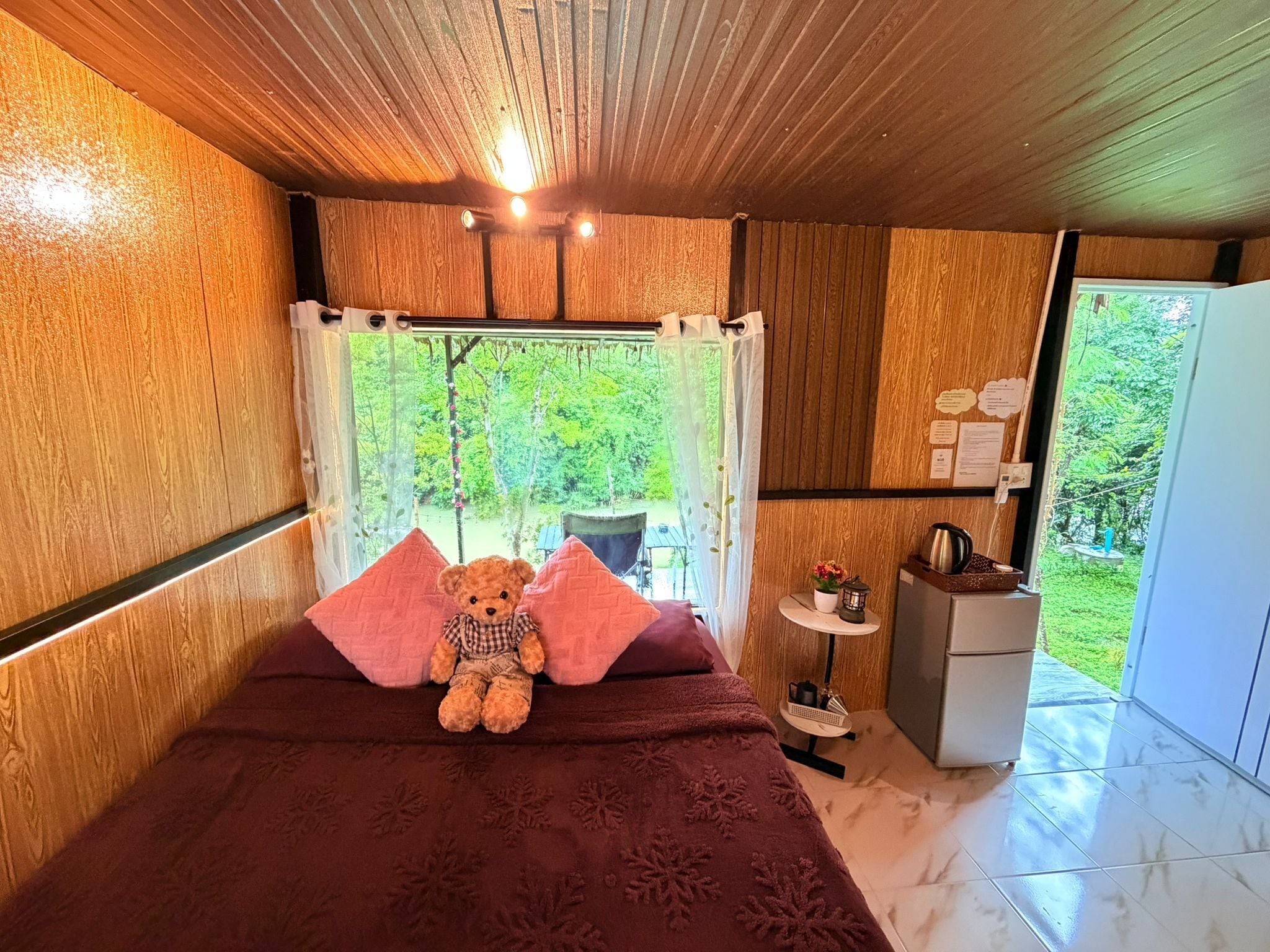 A room with a bed, a teddy bear, a small table with a kettle, and a view of greenery outside.