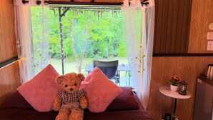 Une pièce chaleureuse avec un lit, un ours en peluche, une fenêtre donnant sur un paysage verdoyant et une petite table sur laquelle est posée une lanterne.