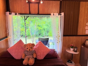 Una habitación acogedora con una cama, un osito de peluche, una ventana con vistas al verdor y una mesita con un farol.