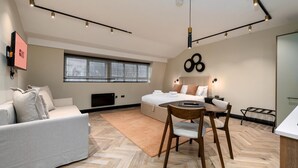 A modern hotel room with a sofa, a dining table with chairs, a bed, a large window, and a wall-mounted artwork. - Guestready - The Euston Hotel (London)