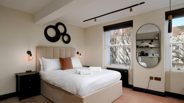 A bedroom with a large bed, bedside tables, a window with a view, a mirror, and a wall-mounted light fixture. - Guestready - The Euston Hotel (London)