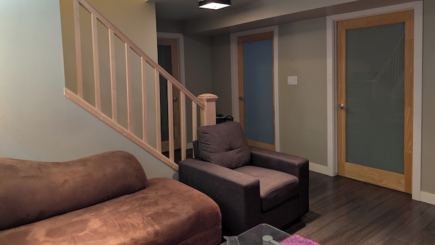 Un salon avec un canapé marron, un fauteuil gris et une table basse en verre. Un escalier en bois, doté d’une rampe en verre, mène à une autre pièce.