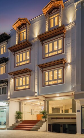Un hotel con fachada moderna, múltiples ventanas con contraventanas de madera y una entrada acogedora con escalones y plantas en macetas.