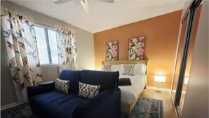 Una habitación de hotel con un sofá azul, una cama con almohadas estampadas, una ventana con cortinas y dos obras de arte en la pared.