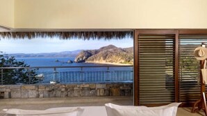 Ein Balkon mit Blick auf einen Strand und das Meer, ein Reetdach und ein Holzstuhl.