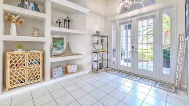 A spacious entryway with white tiled flooring, a wooden cabinet with shoes, shelves with decorative items, and a 'WELCOME' sign.
