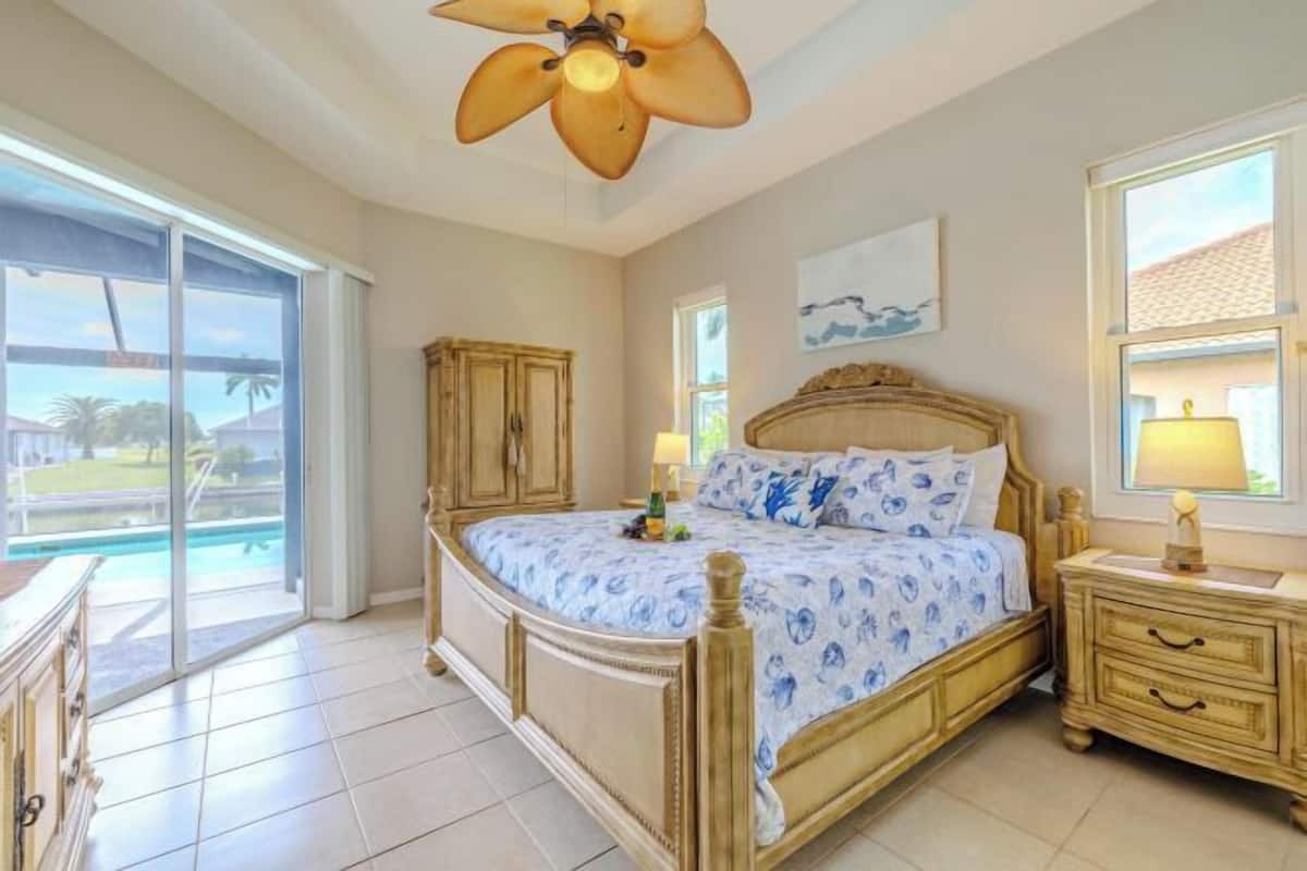 Une chambre à coucher comprenant un lit, une table de chevet, un ventilateur de plafond, une fenêtre et une vue sur une piscine.