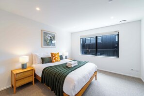 A bedroom with a bed, nightstand, lamp, and a window with a view of a building. - Mountainlight Escape - With Lake and Alpine Views (Wānaka)