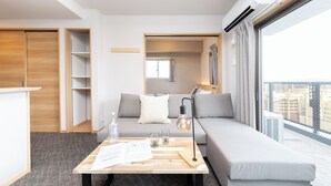 A modern hotel room with a grey sofa, a wooden coffee table with papers, glasses, and a bottle, and a large window offering a city view. - Residence Hotel Hakataekiminami (Fukuoka)