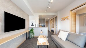 A modern living room with a flat-screen TV, a wooden coffee table, a grey sofa, and a kitchen area in the background. - Residence Hotel Hakataekiminami (Fukuoka)