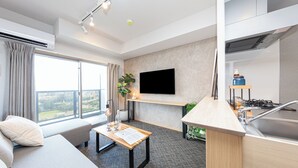 A modern hotel room with a sofa, a small table, a flat-screen TV mounted on the wall, a kitchen area with a sink and stove, and a large window offering a city view. - Residence Hotel Hakataekiminami (Fukuoka)
