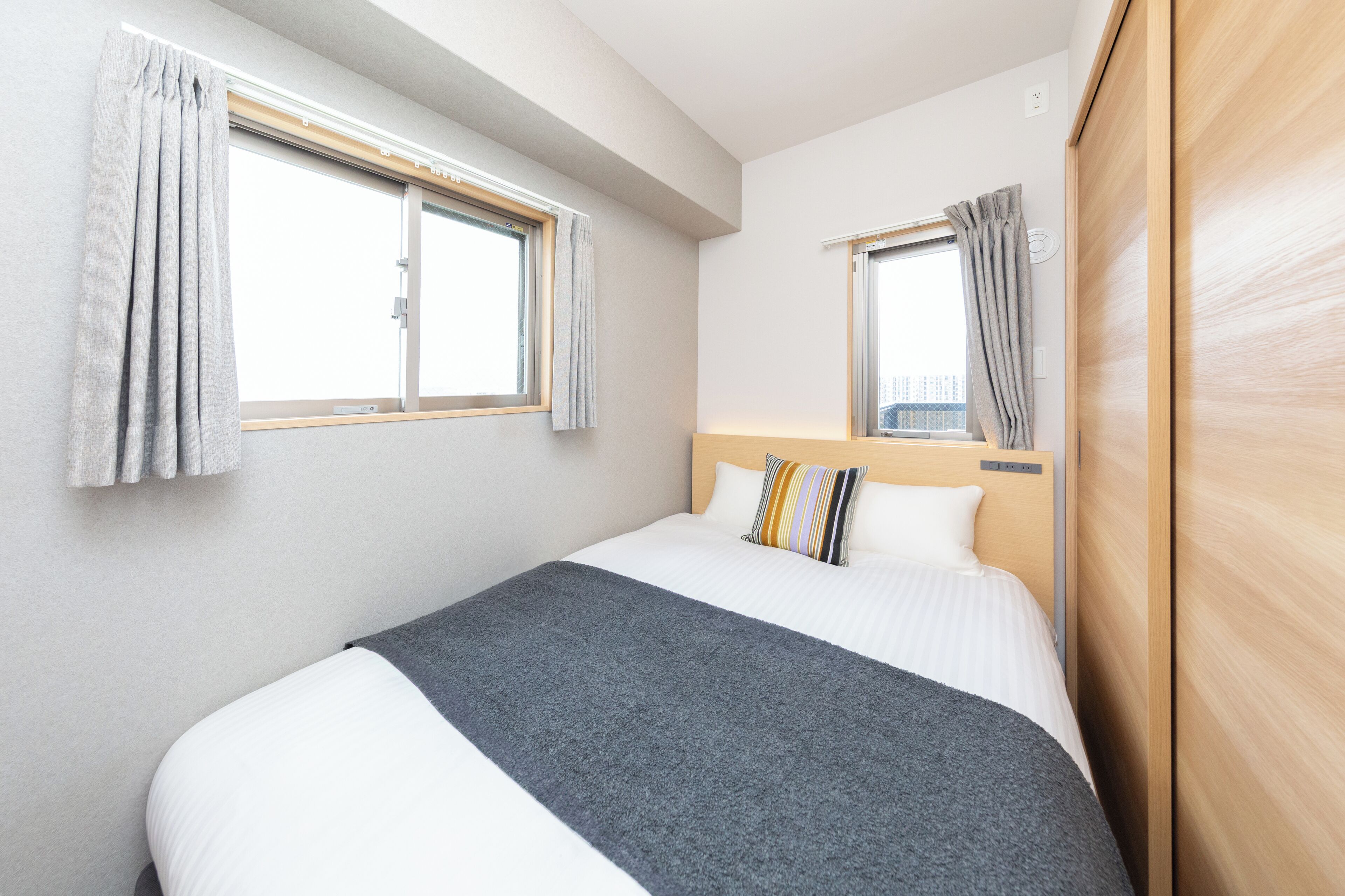 A hotel room with a bed, two windows, and a wooden wardrobe.