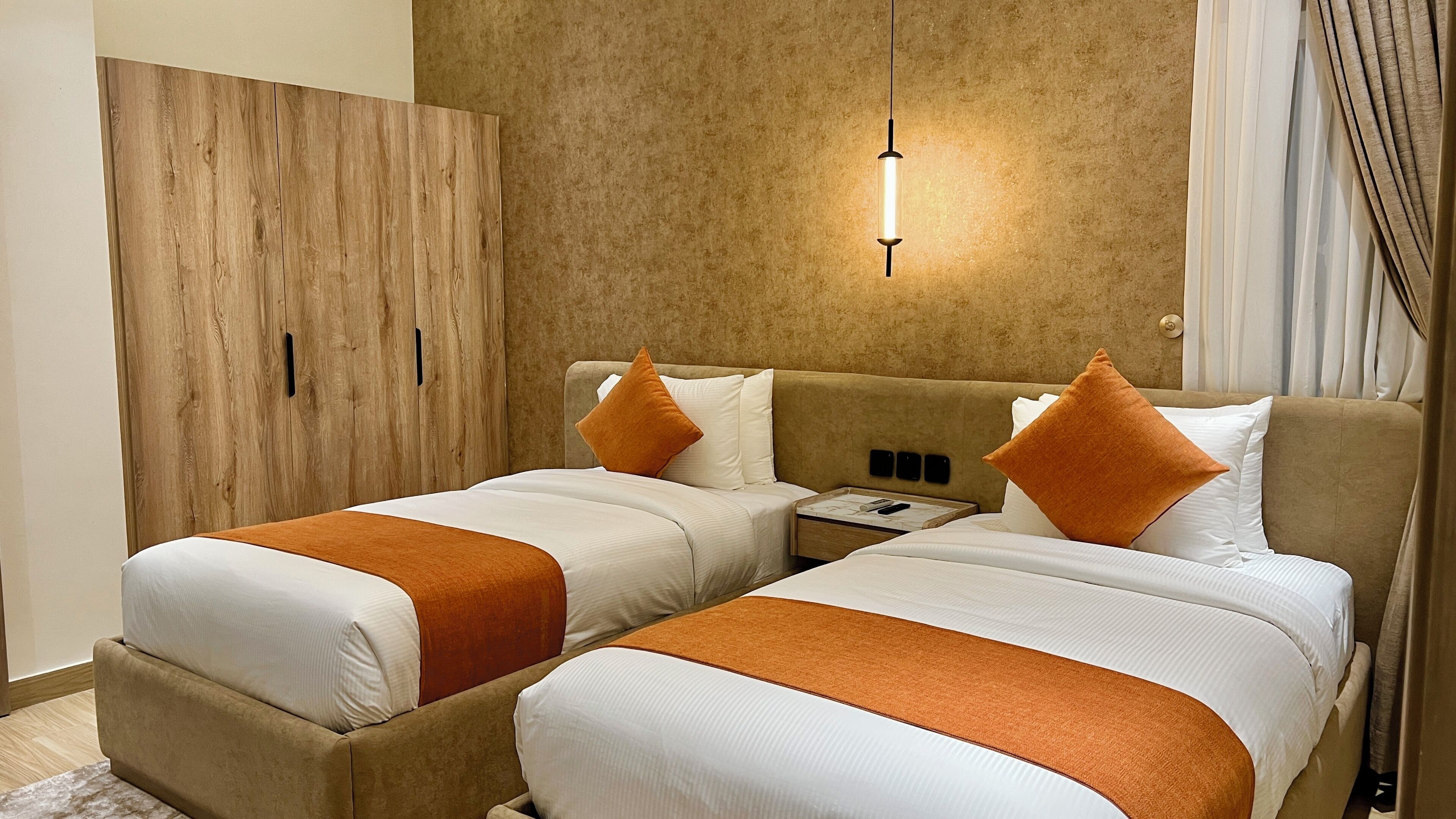 A hotel room with two beds, a wooden wardrobe, a nightstand, and a hanging light fixture.