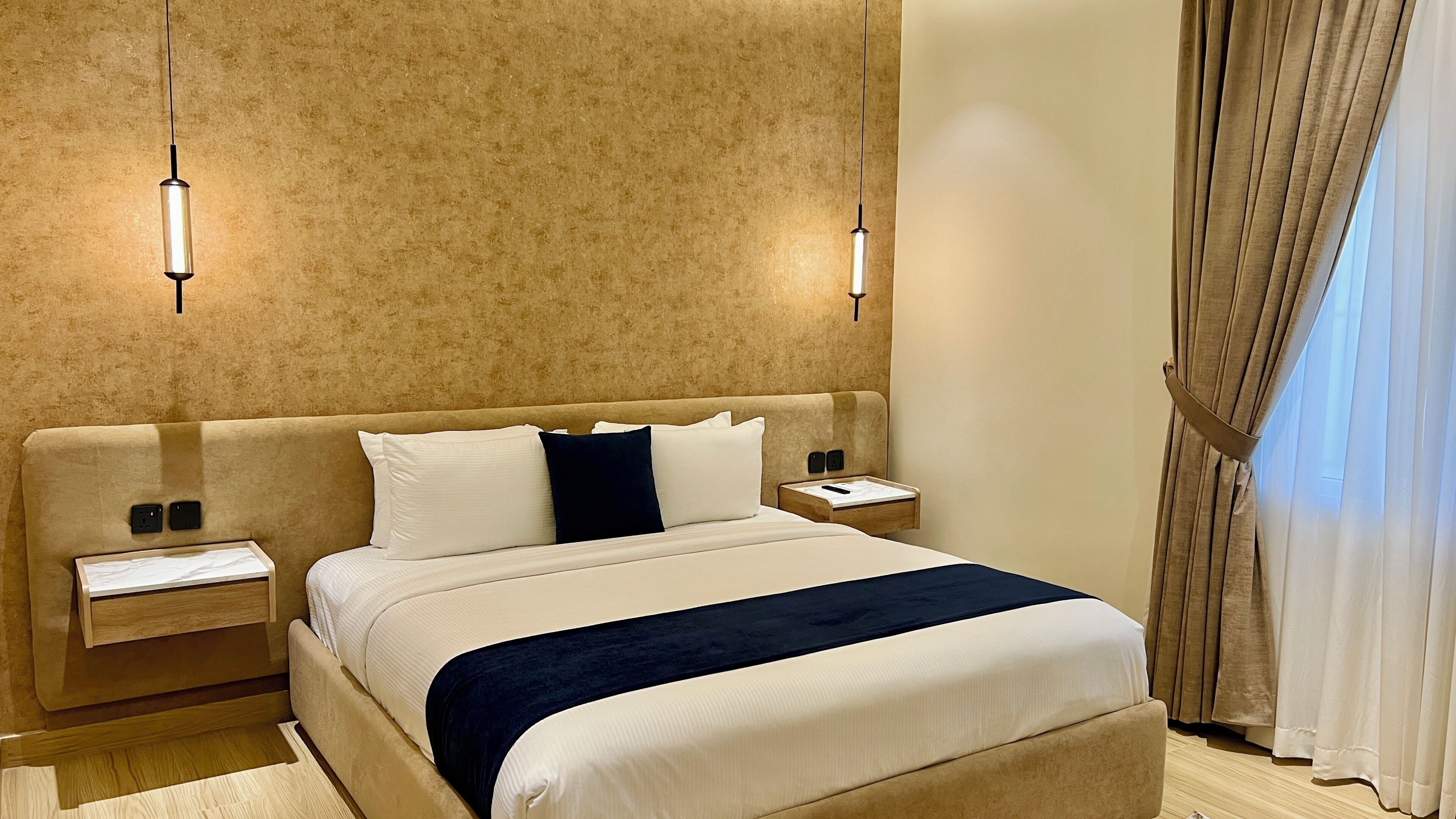 A hotel room with a large bed, bedside tables, a nightstand, a wall-mounted lamp, and a window with curtains.