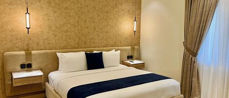 A hotel room with a large bed, bedside tables, a nightstand, a wall-mounted lamp, and a window with curtains.