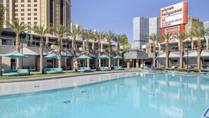 Un espace piscine doté de chaises longues et de parasols, entouré de palmiers et des bâtiments de l’hôtel.