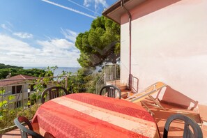 Eine Terrasse mit Tisch, bedeckt mit einer rot-weißen Tischdecke, umgeben von Stühlen und mit Blick auf das Meer und grüne Landschaften.