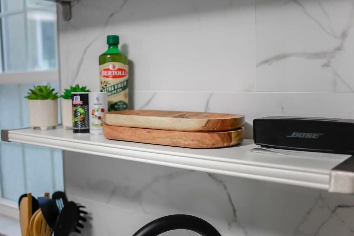 Una repisa en la cocina con una botella de aceite de oliva Bertolli, una lata de café, una caja de té y una tabla de cortar de madera.