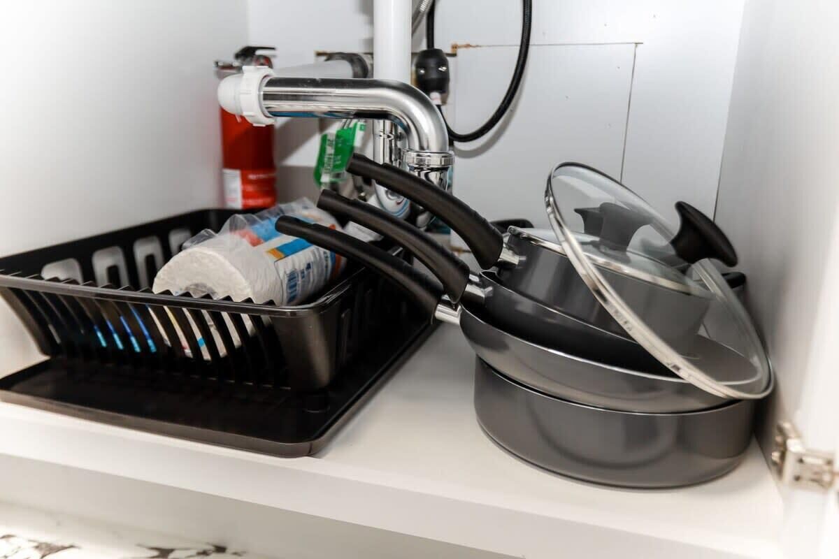 Un lavabo de cocina con un grifo, un escurreplatos negro y una olla con tapa.