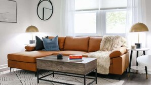 A modern living room with a brown leather sofa, a wooden coffee table, and a white rug. There are two lamps on either side of the sofa, and a framed abstract artwork on the wall. - Sojourn | The South Shore Apartments (Norfolk)