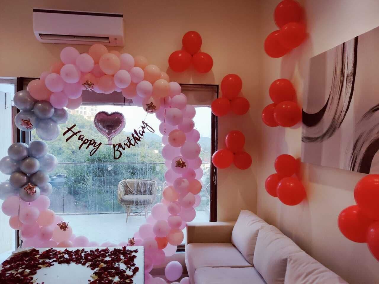 Een kamer met een groot raam, een witte bank en een wand versierd met een ballonboog en een boeket hartvormige ballonnen.
