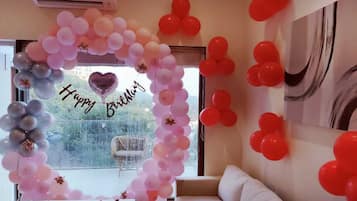 Een kamer met een groot raam, een witte bank en een wand versierd met een ballonboog en een boeket hartvormige ballonnen.