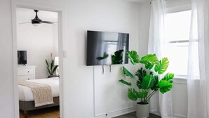 Et moderne soveværelse med en seng, et fjernsyn ophængt på væggen, en loftsvifte og en stor stueplante.