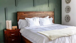 A neatly made bed with white linens and pillows, a wooden nightstand with drawers, and a decorative wall piece. - Sojourn | EastBay Gateway with Game Room (Norfolk)