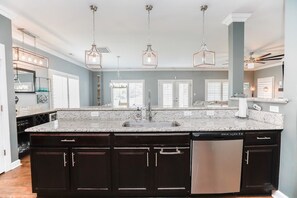 A kitchen with a large island, dark cabinetry, a stainless steel dishwasher, and pendant lighting. - Sojourn Baycation Beach House 5 Min Walk to Beach (Norfolk)