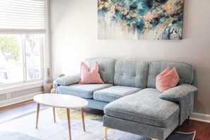 Ein modernes Wohnzimmer mit einem grauen Ecksofa, zwei rosafarbenen Kissen, einem weißen Couchtisch und einem großen abstrakten Gemälde an der Wand.