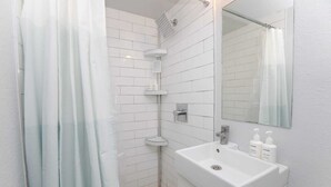 Ein Badezimmer mit weißen Fliesen, einer Dusche mit Glastür, einem Waschbecken mit Spiegel und Pflegeprodukten.