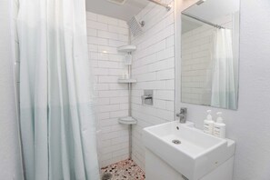 Ein Badezimmer mit weißen Fliesen, einer Dusche mit Glastür, einem Waschbecken mit Spiegel und Pflegeprodukten.