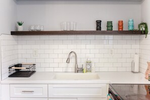 A kitchen with white subway tiles, a stainless steel sink, a wooden shelf with glassware, and a plant. - Sojourn Tide Suite 2 BR 2 BA at Beach Cottages #2 (Norfolk)