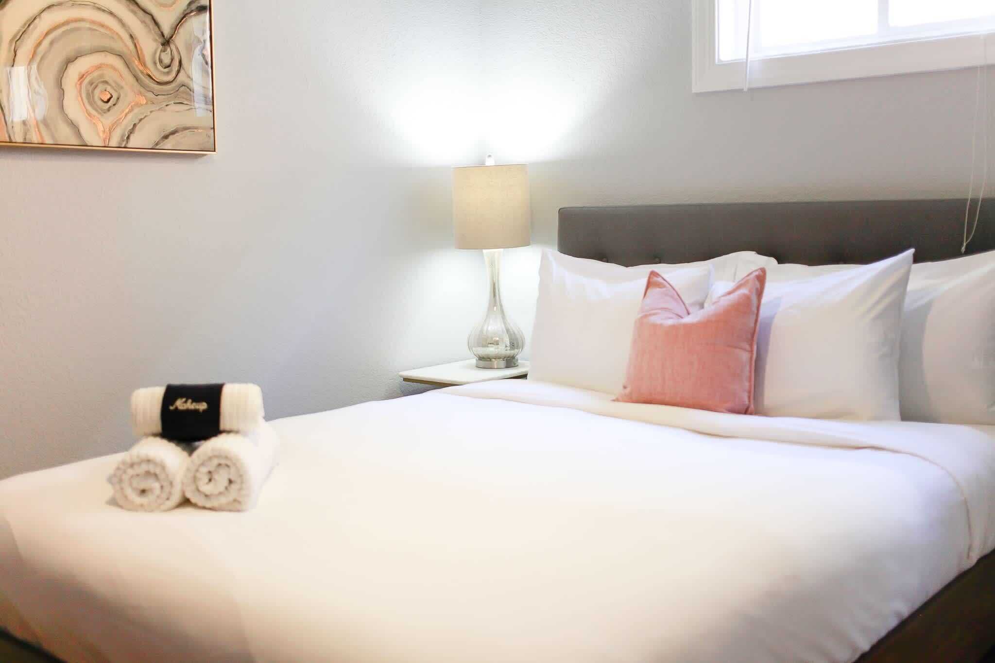 Un lit bien fait, avec du linge de lit blanc, un coussin rose et des serviettes enroulées sur une table de chevet surmontée d’une lampe.