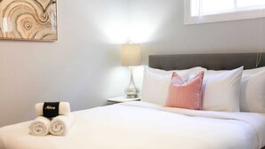 A neatly made bed with white linens, a pink pillow, and rolled towels on a nightstand with a lamp. - Sojourn Cove Suite 2 BR 2 BA at Beach Cottages (Norfolk)