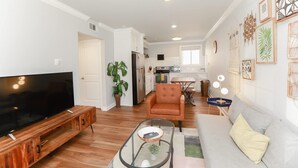 A modern living room with a flat-screen TV, a sofa, a coffee table, and a dining area with a table and chairs. - Sojourn Cove Suite 2 BR 2 BA at Beach Cottages (Norfolk)