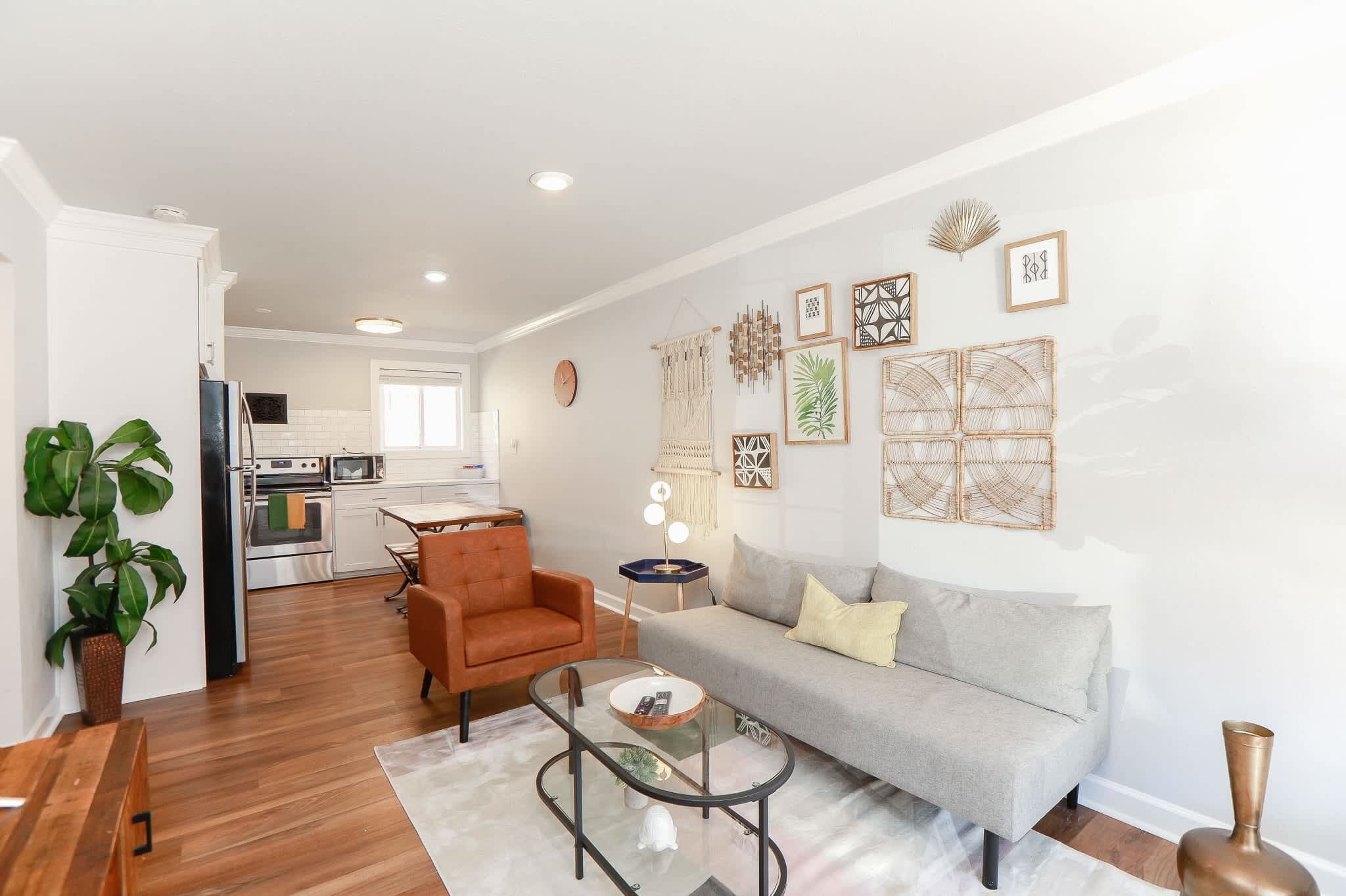 A modern living room with a sofa, armchair, and coffee table, a kitchen in the background, and decorative wall art.